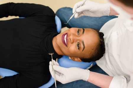 Young African-American ethnic black female smiling while dentist in white latex gloves check condition of her teeth.の写真素材