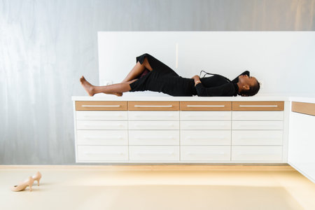 Elegant african or black american woman in dark dress posing on desk in light interior.