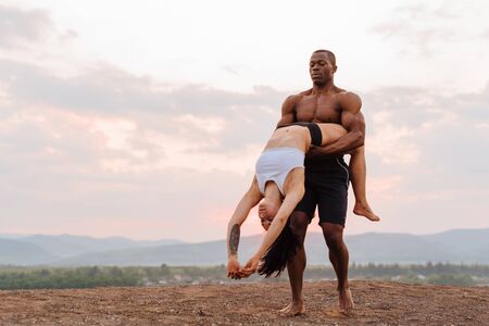 Mixed race gymnastic couple with perfect muscular bodies in sportswear dancing on mountains landscape background. Pink sunset sky and white clouds.の写真素材