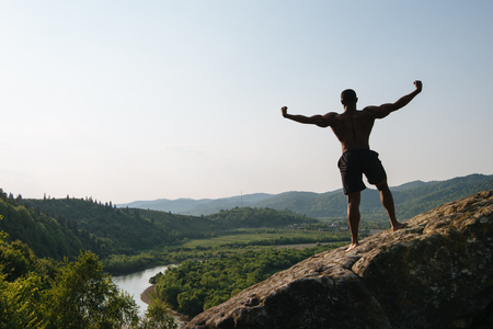 Back view. Silhouette of african american athletic man with naked torso posing on the rock. Green mountain nature background.の写真素材