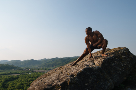 Strong handsome african american bodybuilder is training on the rock in mountains. Green forest mountain background.の写真素材