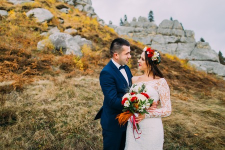 Beautiful wedding couple on idyllic pastoral landscape with rocks and fence as background. Groom in stylish blue suit, white dressed bride holding bouquet of roses.の写真素材