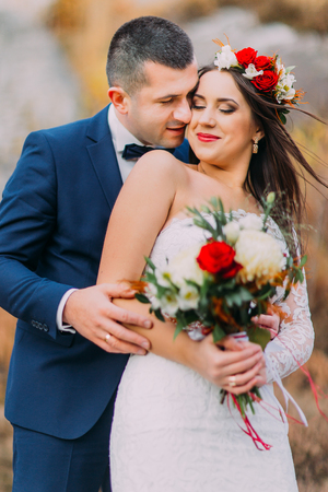 Loving groom kissing his pretty new wife on the cheek in the countryside. Light wind flapping women's hair under stylish head wreath.の写真素材
