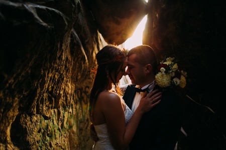 Romantic beautiful bride and elegant stylish groom holding each other in darkened rock cleft backgrounded by sunset shining.の写真素材