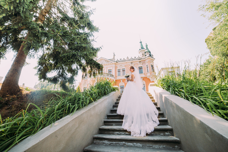 Beautiful bride in magnificent dress going up the stone stairs to antique romantic building at sunny day.の写真素材