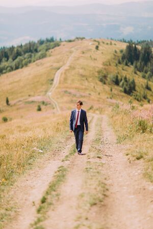 Young man in stylish suit walking on trail by summer field with hills at background.の写真素材