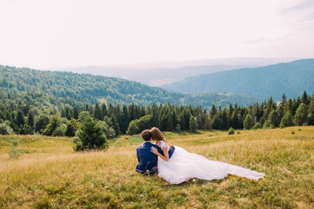 Beautiful bride in hands of her loving groom sitting on yellow grass at sunny field with majestic forest hills as background.の写真素材