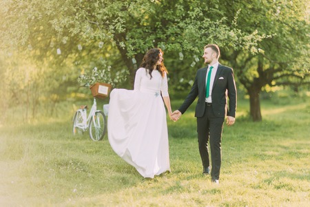 Happy newlyweds walking on grass in park with their bicycle near the tree at background.の写真素材