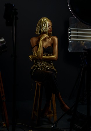 Surreal Portrait of Sexy African American Woman with Glossy Golden Makeup and Headwear Posing To The Camera in Studio. Bronze Bodypaint, Black Studio Background.の写真素材