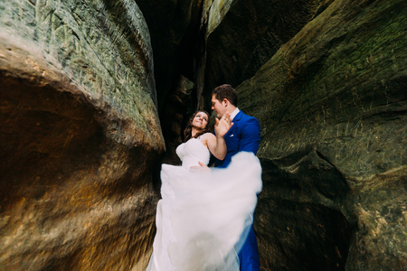 Young romantic bride in white wedding dress with long tail and her loving groom posing at darkened rock cleft.の写真素材