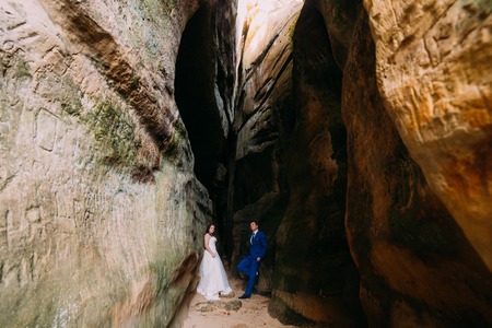 Young romantic bride with long white dress and her groom in stylish blue suit posing at darkened rock cleft.の写真素材