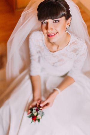 Excited bride in gorgeous white dress waiting for her wedding posing with cute floral boutonniere sitting in dressing room before the ceremony.の写真素材