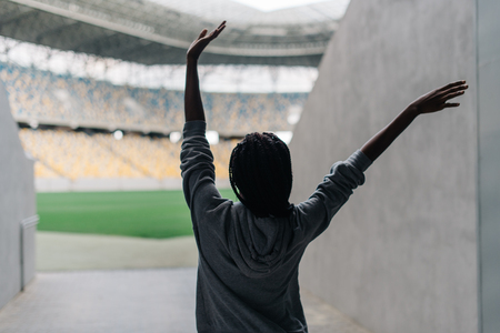 Happy african american woman standing among empty stadium celebrating with her arms raised punching the air with her hands, view from back.の写真素材