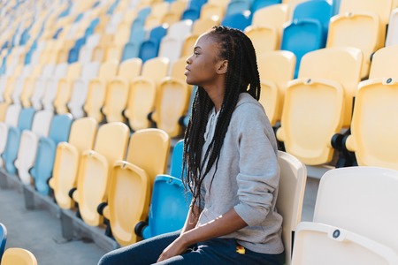 Excited african american woman in gray sportswear looking away sitting on bleachers waiting for a show, celebration.の写真素材