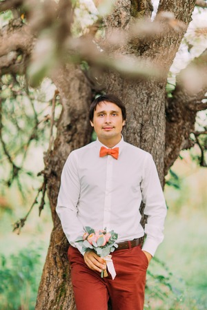 Handsome confident groom with a bouquet of flowers looking at camera leaning on tree in summer park, bright sunny dayの写真素材