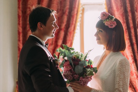 Happy luxury bride and groom looking at each other standing at window light in room, tender moment.の写真素材