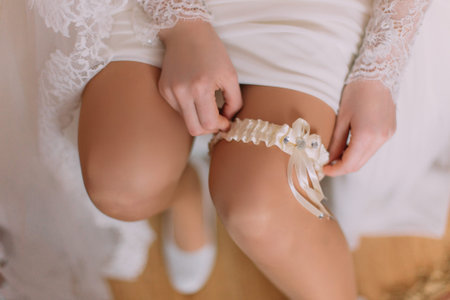 Beautiful bride in dressing room is wearing bridal garter, preparing herself for wedding ceremony. Close-up arms of woman.の写真素材