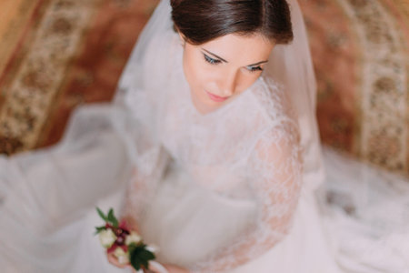 Close-up portrait of beautiful innocent bride in wedding dress sitting on carpet and holding cute floral boutonniere.の写真素材