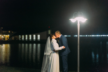 Romantic wedding. Cheerful bride and groom kissing near night lake illuminated with bright light from banquet hall windows.の写真素材