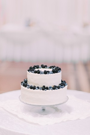 Candid cream cake decorated with bilberries and blackberries isolated close-up on table covered by white tablecloth at blurred bright background of restaurant interior.の写真素材