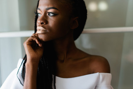portrait photo of black girl in white dress with bare shoulders. she put her hand to her beautiful face and looking at somewhere. evening make upの写真素材