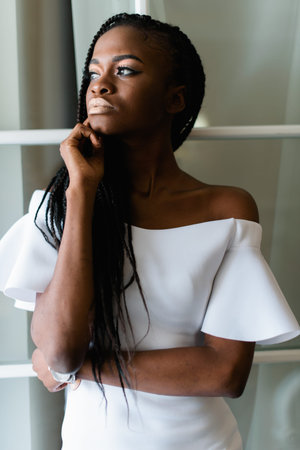 attractive sexy black woman standing near window in white dress with bare shoulders. she put her...