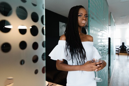 beautiful african woman in white dress with bare shoulders drinking a glass of red wine.の写真素材