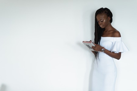 Sexy pretty photo model with black skin is standing in front of a white wall and chatting in internet using her tablet. Girl is wearing long white dress with bare shoulders and watch on her handの写真素材