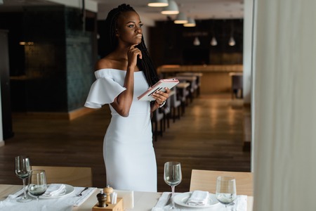 Adorable sexy woman with black skin is stending in luxury restaurant near well decorated table. Girl has tablet in her hand. Model is looking at the window and thinking about something.の写真素材