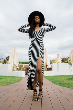 Charming, pretty, tender, beautiful african woman wearing dress in stripes and black hat on her head. Model staying on luxury hotel terraceの写真素材