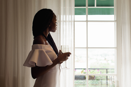 Charming, touching, tender, beautiful african woman in restaurant staying in front of the big window. Girl is in whihe dress, has long dark hair and a glass of alcohol in her hands.の写真素材