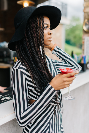 Girl with dark skin in bar with red cocktail in her hand. Model put her another hand on her hat. Ice cocktails. Margarita.の写真素材
