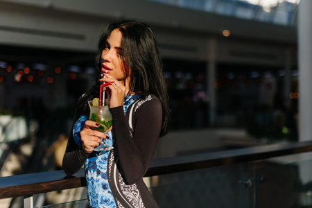 Attractive brunette with cocktail in her hand staying in the shopping mall. She wearing elegant dress. Look at the camera. Mohito with ice and mintの写真素材
