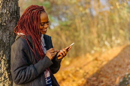 Pretty woman standing near tree in the autumn park at sunset. Girl chatting in internet using her phone and smiling. Beautiful autamn parkの写真素材