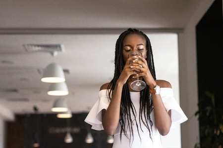 Beautifull dark-skinned girl drinking wine from a glass in a restaurant. Dreadlocks or african braides on her head.の写真素材