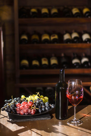 Bottle of wine, wine glass and plate with grapes placed on a wooden table in a wine vault.