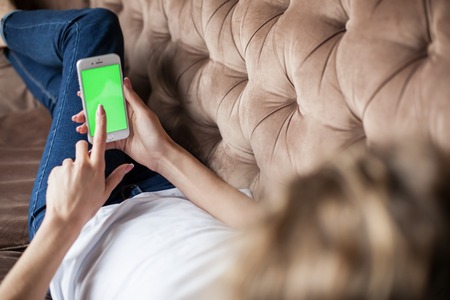 Beautiful girl lying on the sofa with the phone in her hands. Phone with green screen.の写真素材