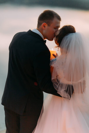Kissing newlywed couple is holding the shining candle at the background of the river.