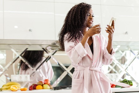 Attractive black woman standing in the kitchen with the phone and glass of water in her hands. Woman wearing pink bathrobe.の写真素材