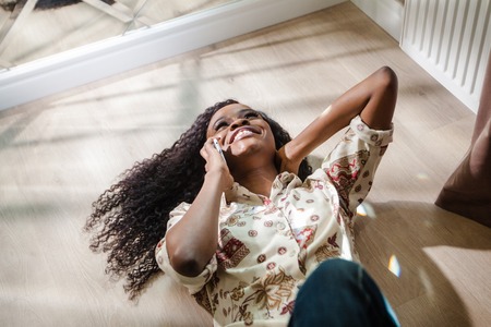 Happy black woman lying on the floor and talking on the phone. Woman with long dark hair.の写真素材