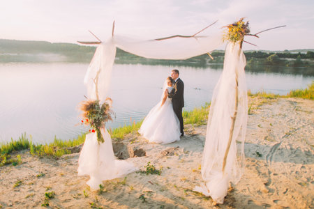 The horizontal view of the hugging newlyweds enjoying the vacation near the decorated arch and river.の写真素材