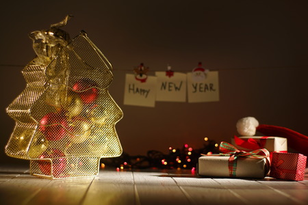 The golden box for balls in the form of the Christmas tree is placed near the wrapped presents at the blurred background of the glowing garlands and new year signsの写真素材