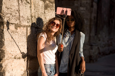 Two attractive girls wearing casual closes and glasses making selfie near the wall. One girl is...