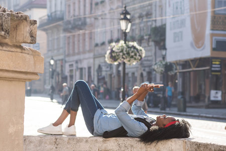 Attractive young black woman lying near the monument in the center of the city with the tablet in her hands. Woman wearing casual clothes.の写真素材