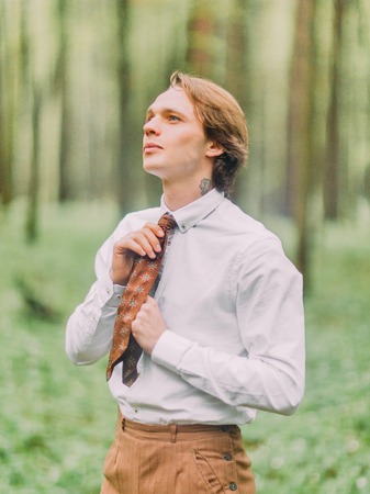 The man with blond hair and tattoo on the neck in the white shirt ties his brown tie and looking at the sky in the green forest. Side portrait.の写真素材