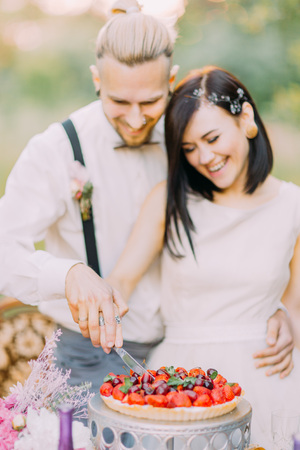 The close-up portrait of the blurred newlywed couple cutting the wedding cake with cherries and strawberries. The wedding dinner is in the sunny field.の写真素材