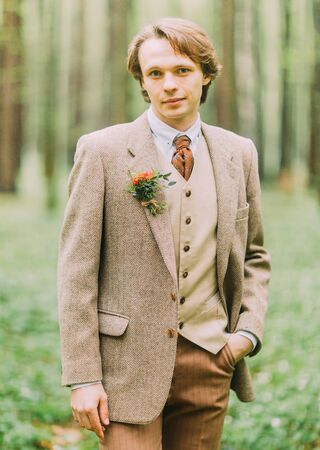Man in the vintage suit, with mini-bouquet of flowers is pinned on his jacket is looking at the camera and keeping hand in his pocket. The close-up front portrait in the wood.の写真素材