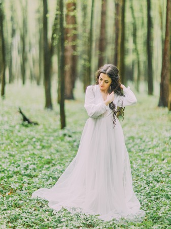 The close-up portrait of the beautiful woman in the white wedding dress looking at the ground and sorting her hair in the green forest.の写真素材