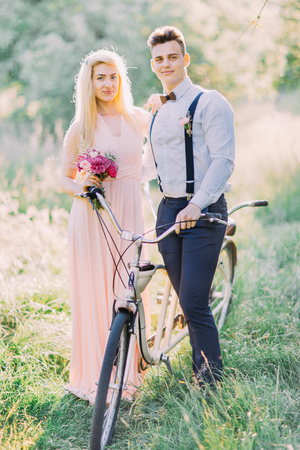 The cute vertical portrait of the bridesmaid holding the pink bouquet and the best man in the blue suit carrying the bicycle in the sunny green wood.の写真素材