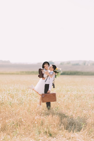 The groom with the vintage suitcase is holding the smiling bride in the arms at the background of the sunny field.の写真素材